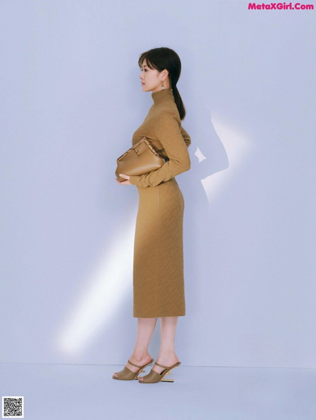 A woman in a brown dress standing in front of a white wall.