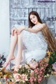 A woman in a white dress sitting on a wooden bench surrounded by flowers.